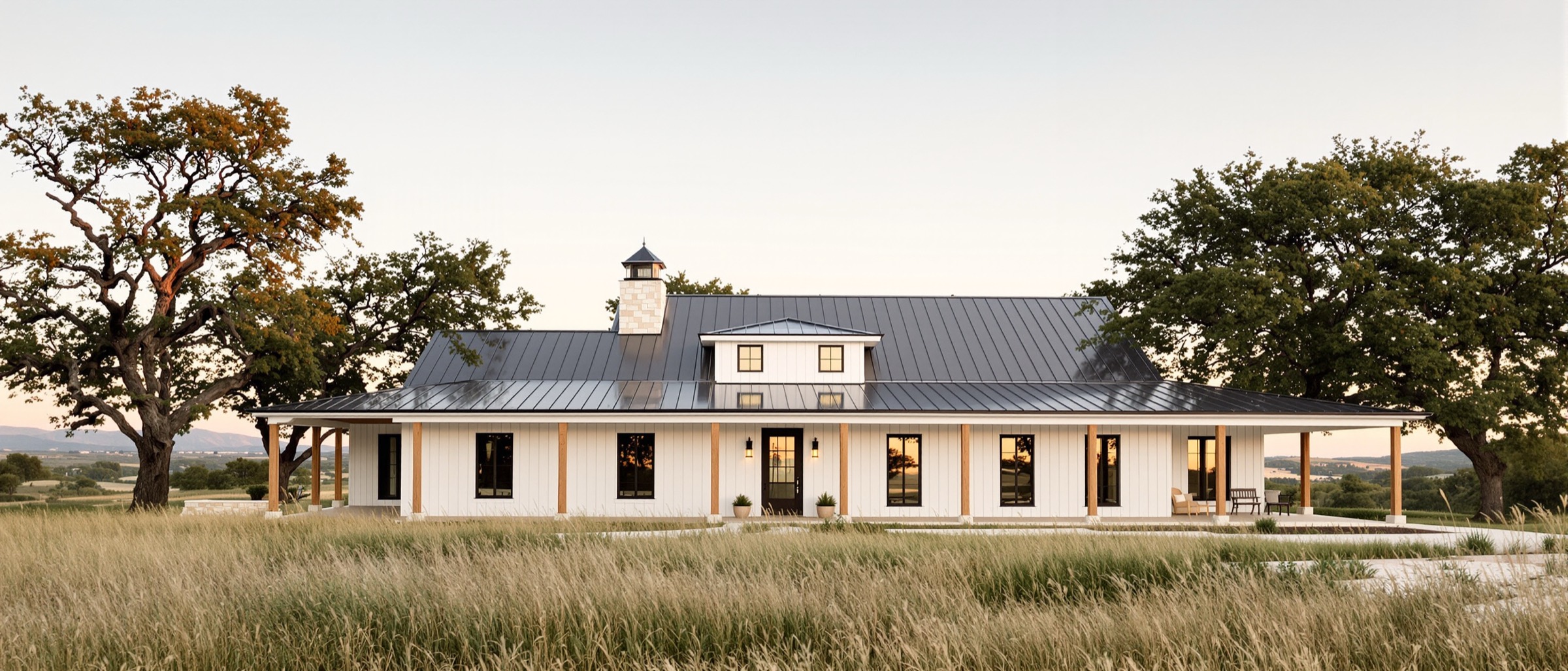 Single-story barndominium with white board-and-batten siding, a charcoal standing seam roof, and a long cedar-post porch among live oaks